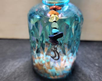 May include: A turquoise glass bottle with a cork stopper, filled with small, colorful stones. A chain of colorful gemstones hangs from the cork, ending with a silver crescent moon charm. The bottle has a textured surface.