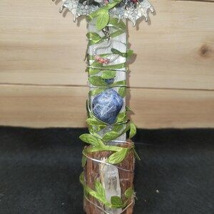 May include: A vertical selenite crystal wand wrapped in silver wire and green fabric leaves. A silver dragon pendant with red accents sits at the top. A blue sodalite stone is attached to the center, and a clear quartz point is at the base.