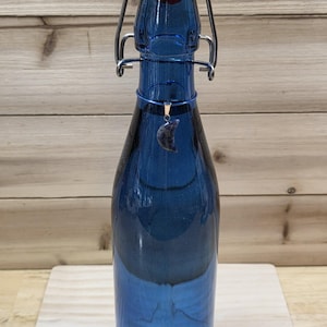 May include: A cobalt blue glass bottle with a swing-top closure featuring a white stopper and a red rubber gasket. A small, crescent moon-shaped pendant with a mottled grey and blue stone finish hangs from a thin wire around the bottle neck.