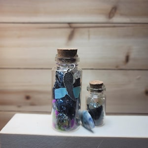 May include: Two small glass bottles with cork stoppers. The larger bottle contains colorful crystals and a silver wing charm. The smaller bottle holds dark stones. A blue and white stone sits in front of the bottles.