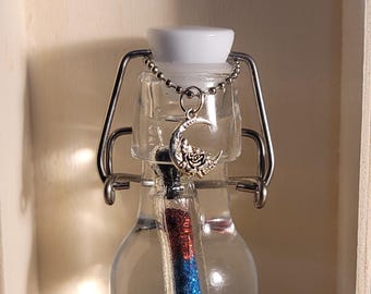 May include: A small glass bottle with a silver metal clasp and a silver crescent moon charm. The bottle is filled with a clear liquid and contains a red, white, and blue glitter stick. The bottle is sitting on a wooden surface.
