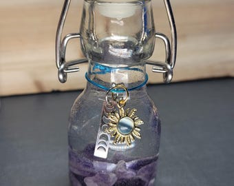 May include: A clear glass bottle with a white stopper and a metal clasp. The bottle contains purple amethyst stones and is adorned with a charm featuring a sun and crescent moons. A blue string secures the charm to the bottle.