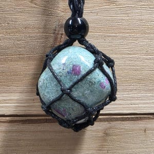 May include: A polished, light green stone with embedded magenta inclusions, secured within a black macrame cord netting. The pendant features a single black bead at the top, attached to a braided black cord necklace for a secure, adjustable fit.