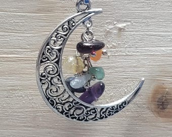 May include: A silver-colored crescent moon pendant with an intricate filigree design. The pendant hangs from a black cord and features a series of colorful gemstones in shades of purple, green, yellow, and orange.