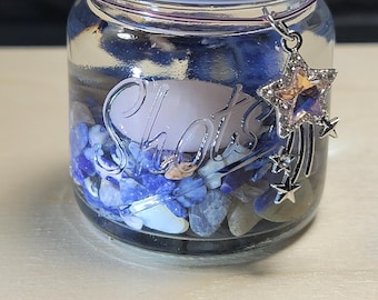 May include: A clear glass jar with a copper-colored lid, filled with blue and white stones. The jar has the word "Shots" on the front and a star charm hanging from the rim. The charm has a large, sparkly star and several smaller stars.