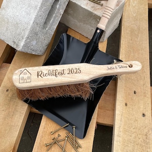 Könnte beinhalten: Eine Holzbürste mit dem Text "Richtfest 2025" und einer Hausgrafik, die auf einer schwarzen Kehrschaufel liegt. Die Bürste hat einen Naturborstenkopf und einen hellen Holzgriff. Der Text "Julia & Tobias" steht ebenfalls auf der Bürste.