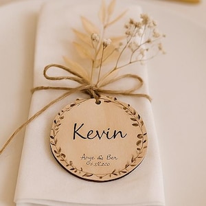 Könnte beinhalten: Tischdekoration für eine Hochzeit. Ein weißer Teller mit Serviette und goldenem Besteck. Ein Namensschild aus Holz mit dem Namen "Kevin" ist mit Bindfaden an der Serviette befestigt und mit einem floralen Design verziert.