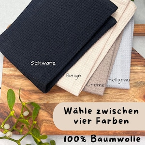 May include: A stack of four waffle-weave towels in black, beige, cream, and light gray. The black towel is folded on top. Text on the image reads "Schwarz", "Beige", "Creme", "Hellgrau", "W&auml;hle zwischen vier Farben", and "100% Baumwolle".
