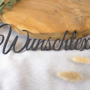 Könnte beinhalten: Ein dunkelgraues Holzschild mit dem Wort "Wunschtext" in Schreibschrift. Das Schild liegt auf einer weißen Stoffoberfläche mit einem Holzschneidebrett im Hintergrund. Trockenblumen und Grünzeug sind ebenfalls vorhanden.