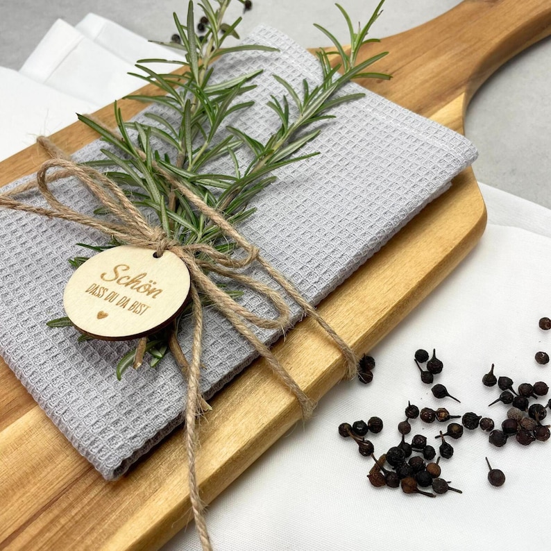 May include: A wooden cutting board with a gray waffle-weave towel and fresh rosemary sprigs tied with twine. A small wooden tag with text is attached. Scattered peppercorns are in the background.