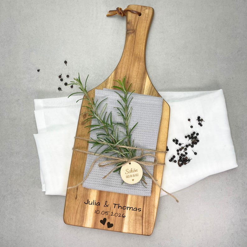 May include: Wooden cutting board with a handle, featuring a gray towel, rosemary sprigs, and a small wooden tag. The board is engraved with names, a date, and two hearts. A white cloth and peppercorns are also present.