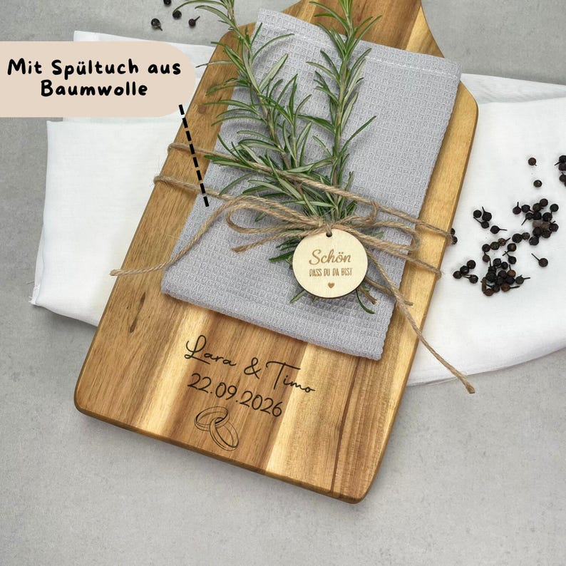 May include: Wooden cutting board with a gray cloth, rosemary sprigs, and a wooden tag tied with twine. The tag reads "Sch&ouml;n" and "dass du da bist". The board is engraved with names, a date, and wedding rings.