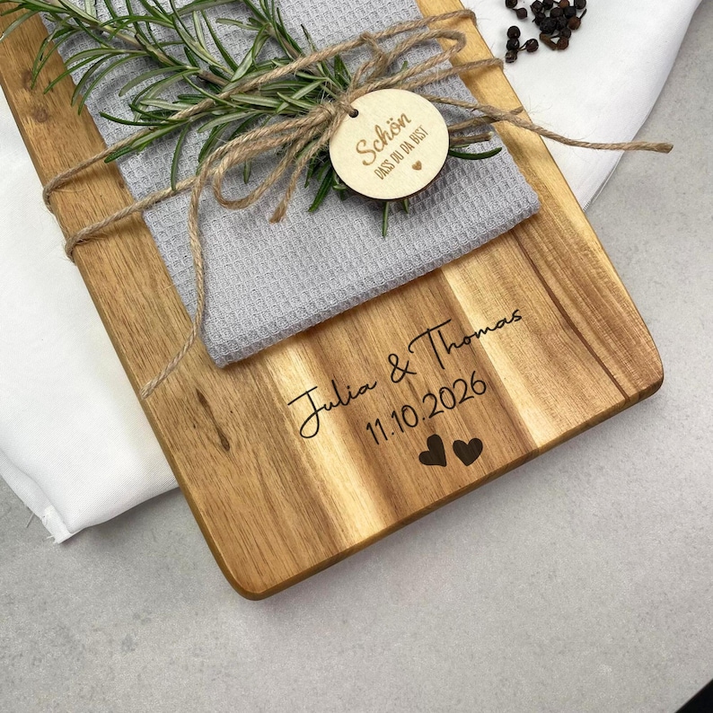 May include: Wooden cutting board with a gray waffle-weave towel, tied with twine and rosemary sprigs. The board is engraved with the names "Julia & Thomas" and the date "11.10.2026", along with two heart symbols. A small wooden tag says "Sch&ouml;n dass du da bist".