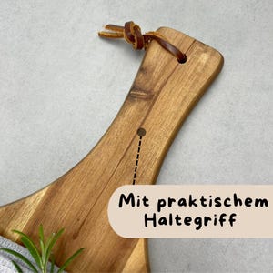 May include: Wooden cutting board with a curved handle and a leather loop for hanging. The board has a natural wood grain and a hole near the handle. The text "Mit praktischem Haltegriff" is visible on the bottom right.