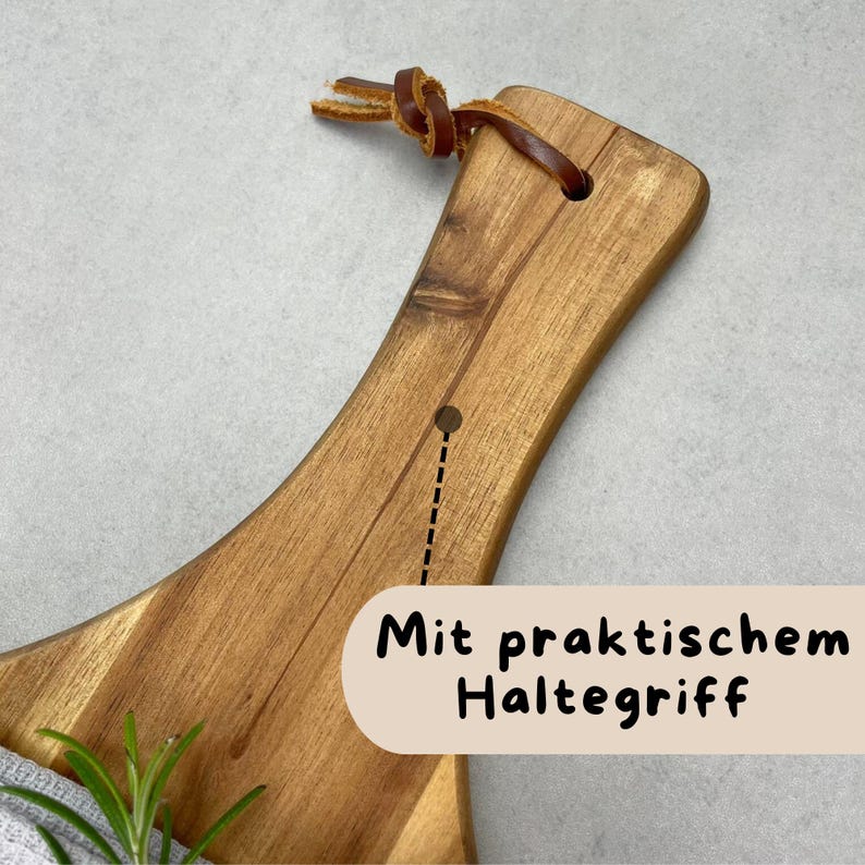 May include: Wooden cutting board with a curved handle and a leather loop for hanging. The board has a natural wood grain and a hole near the handle. The text "Mit praktischem Haltegriff" is visible on the bottom right.