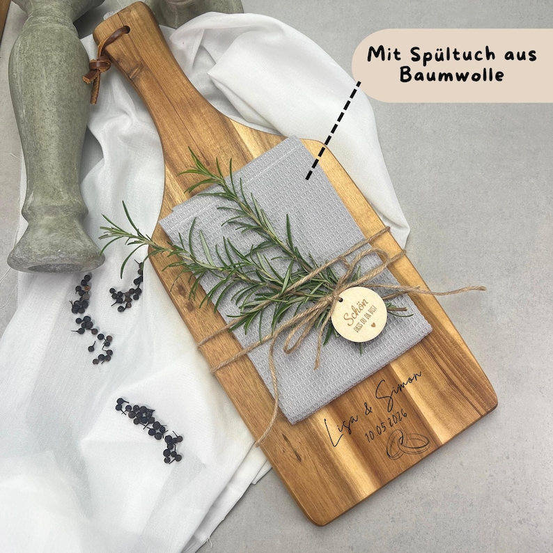 May include: A wooden cutting board with a handle, holding a gray washcloth and rosemary sprigs, tied with twine. A small wooden tag and the names "Lisa & Simon" with a date and rings are engraved on the board. The text "Mit Sp&uuml;ltuch aus Baumwolle" is also present.