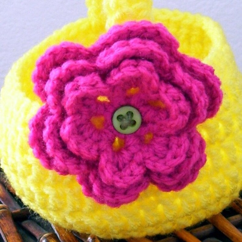May include: A yellow crocheted flower pot with a pink crocheted flower on top. The flower has a green button in the center.