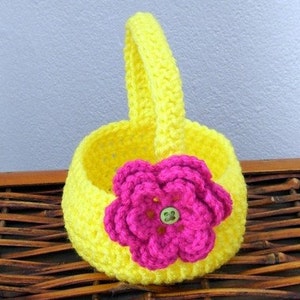 May include: A small yellow crocheted basket with a pink crocheted flower and a green button in the center. The basket has a handle and is sitting on a woven brown surface.