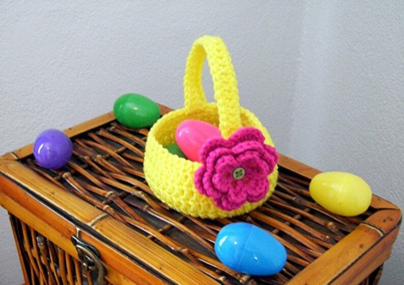 May include: A yellow crocheted Easter basket with a pink crocheted flower and a green button in the center. The basket is filled with plastic Easter eggs in pink, blue, and green.