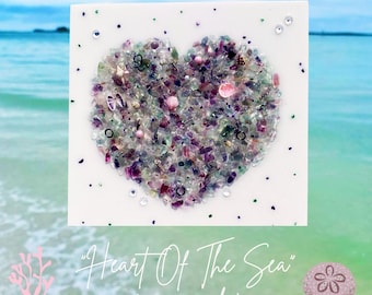 May include: A white card with a heart-shaped design made of colorful sea glass and other small objects. The text "Heart Of The Sea" (mermaid) is written below the heart.
