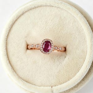 May include: A rose gold ring featuring a central oval-cut deep pink gemstone surrounded by a halo of small white stones. The band is crafted from rose gold and includes additional small white stones set along the shoulders of the ring.