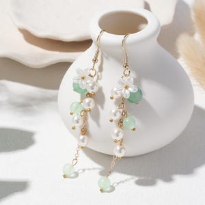 Peut inclure: Boucles d'oreilles pendantes élégantes avec des fleurs blanches, des perles vert clair et des fausses perles. Les boucles d'oreilles ont des crochets et des chaînes dorés. Les boucles d'oreilles sont présentées sur un vase blanc.
