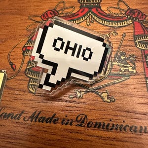 May include: A white and black pixelated speech bubble with the word "OHIO" in black letters. The speech bubble is on a wooden surface with a decorative design.