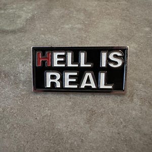 May include: A rectangular pin with the words "HELL IS REAL" in white capital letters on a black background. The word "HELL" has red lettering. The pin has a silver border and is set against a gray surface.