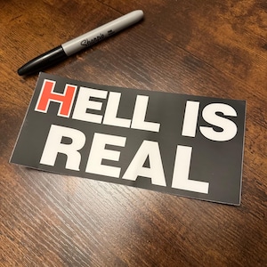 May include: A black sticker with the words "HELL IS REAL" in white letters, with the word "HELL" in red. A silver and black Sharpie pen is next to the sticker on a wooden surface.