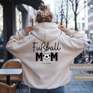 Könnte beinhalten: Ein cremefarbener Kapuzenpullover mit den Worten "Fußball MOM" in schwarzer Schrift und Blockbuchstaben, mit einem Fußballherz-Design. Der Text "VON CRISTIANO" steht darunter. Der Hoodie hat eine Kapuze mit Kordelzug und eine Kängurutasche.