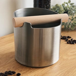 Könnte beinhalten: Ein Edelstahl-Kaffeeabschlagbehälter mit einer Holzstange. Der zylindrische Behälter hat eine gebogene Oberseite und eine Holzstange zum Ausklopfen von Espressopucks. Kaffeebohnen sind auf der Holzoberfläche verstreut.