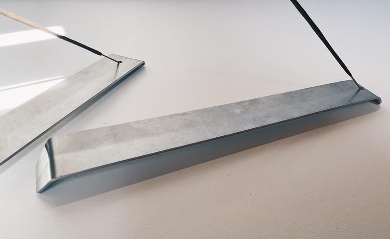 May include: Two silver metal incense holders with a black incense stick resting on one of the holders.
