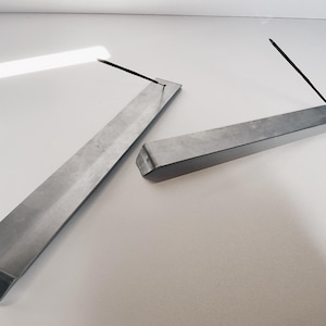 May include: Two silver metal incense holders, each with a rectangular shape and a slight bend at one end. The holders are designed to hold incense sticks.
