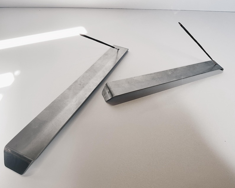 May include: Two silver metal incense holders, each with a rectangular shape and a slight bend at one end. The holders are designed to hold incense sticks.