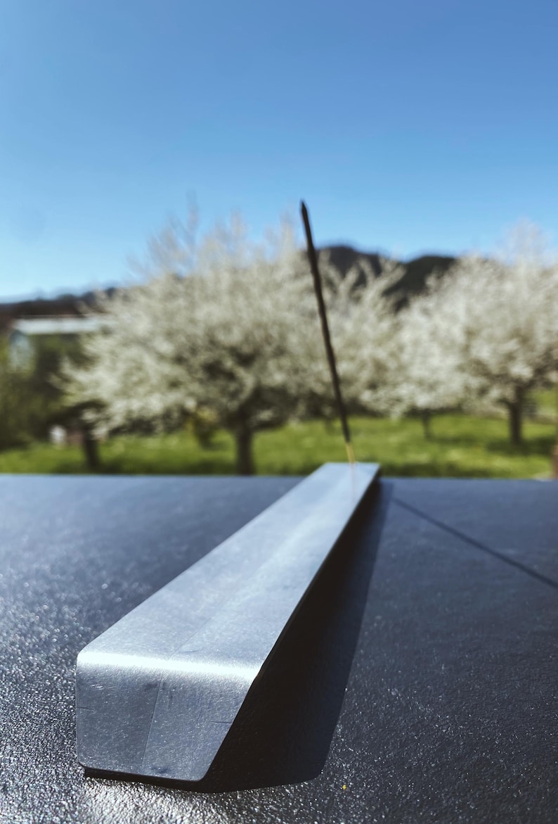 May include: A silver metal incense holder with a black incense stick burning. The holder is on a dark surface with a blurred background of trees with white blossoms.
