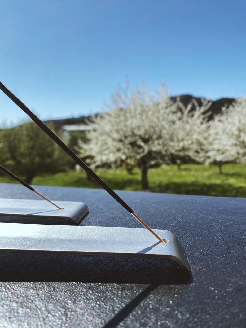 May include: Two silver incense holders with a black incense stick burning on one of them. The holders are on a dark surface with a blurred background of a green field and a white flowering tree.