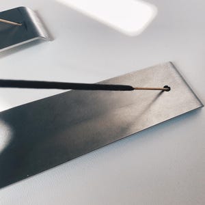 May include: A silver metal incense holder with a black incense stick resting in a small hole. The holder is rectangular and has a smooth surface.