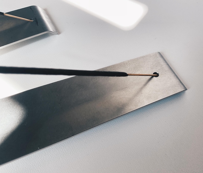 May include: A silver metal incense holder with a black incense stick resting in a small hole. The holder is rectangular and has a smooth surface.