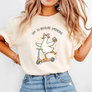 May include: A cream-colored t-shirt featuring a graphic of a white duck wearing a pink bow, riding a pink scooter, and holding a coffee cup. The text above the duck reads "OFF TO REGULATE EMOTIONS" in black, accompanied by small yellow star icons.