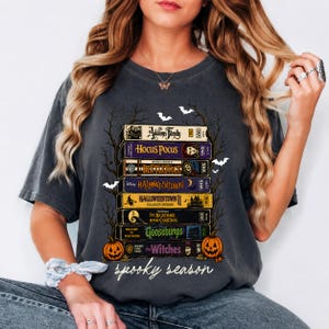 Puede incluir: Una camiseta gris carbón con un estampado gráfico de una pila de cintas VHS etiquetadas como The Addams Family, Hocus Pocus, Beetlejuice, Halloweentown, Halloweentown II, The Nightmare Before Christmas, Goosebumps y The Witches, con el texto spooky season.