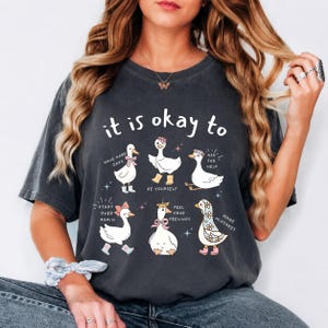 Può includere: Una t-shirt grigio antracite a maniche corte con una grafica di sei oche bianche e il testo "it is okay to" insieme alle frasi "have hard days," "be yourself," "ask for help," "start over again," "feel your feelings," e "make mistakes."