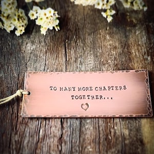 May include: A copper-colored rectangular tag with the words "TO MANY MORE CHAPTERS TOGETHER..." and a heart symbol. The tag is on a wooden surface with white flowers.
