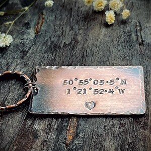 May include: A copper-toned rectangular keyring with engraved coordinates and a heart symbol. The keyring is attached to a textured ring. The background is a dark wooden surface with small white flowers.