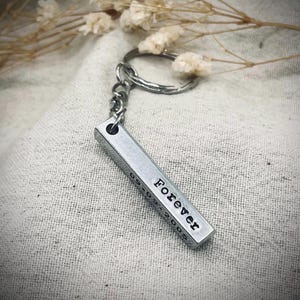 May include: A silver-toned metal bar keychain featuring a split ring attachment. The rectangular bar is engraved with the word Forever and the date 09-09-2005 in black lettering on its side, showcasing a clean, metallic finish and a sturdy design.