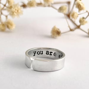 May include: A silver ring with the words "you are enough" engraved on the outside. The ring has a flat, rectangular design with a small gap. The background features soft, blurred white and beige elements.