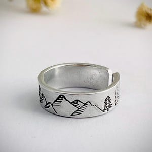 May include: A silver adjustable ring with a mountain and tree design. The ring has a detailed black outline of a mountain range and trees around the band. The ring is made of a shiny, metallic material.