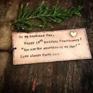May include: A copper-colored metal tag with a heart-shaped accent and the text "To my husband Dan, Happy 10th Wedding Anniversary! You are the sunshine of my life! Love always Sarah." A sprig of green foliage is in the background.