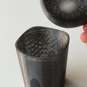 以下が含まれることがあります： 暗い灰色の、テクスチャ加工された容器と、それに合う蓋。容器は円筒形で、上部がわずかに広がっています。蓋は楕円形で、手で持たれています。両方の部品の内側には、より明るい渦巻き模様があります。