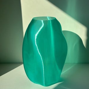 Puede incluir: Un jarrón translúcido de color verde azulado, con una forma orgánica y fluida. El jarrón tiene una abertura ancha y una superficie lisa y brillante. La luz crea reflejos y sombras, enfatizando las curvas. Es un objeto decorativo.