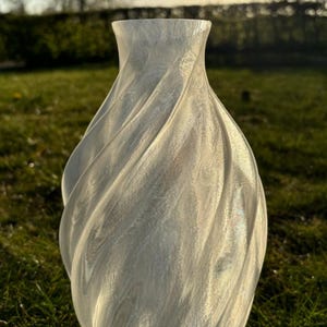 May include: A white, textured vase with a swirling design. The vase has a wide base that tapers to a narrow neck. The vase is made of a translucent material, and the light shines through it. The vase is sitting on a grassy surface.